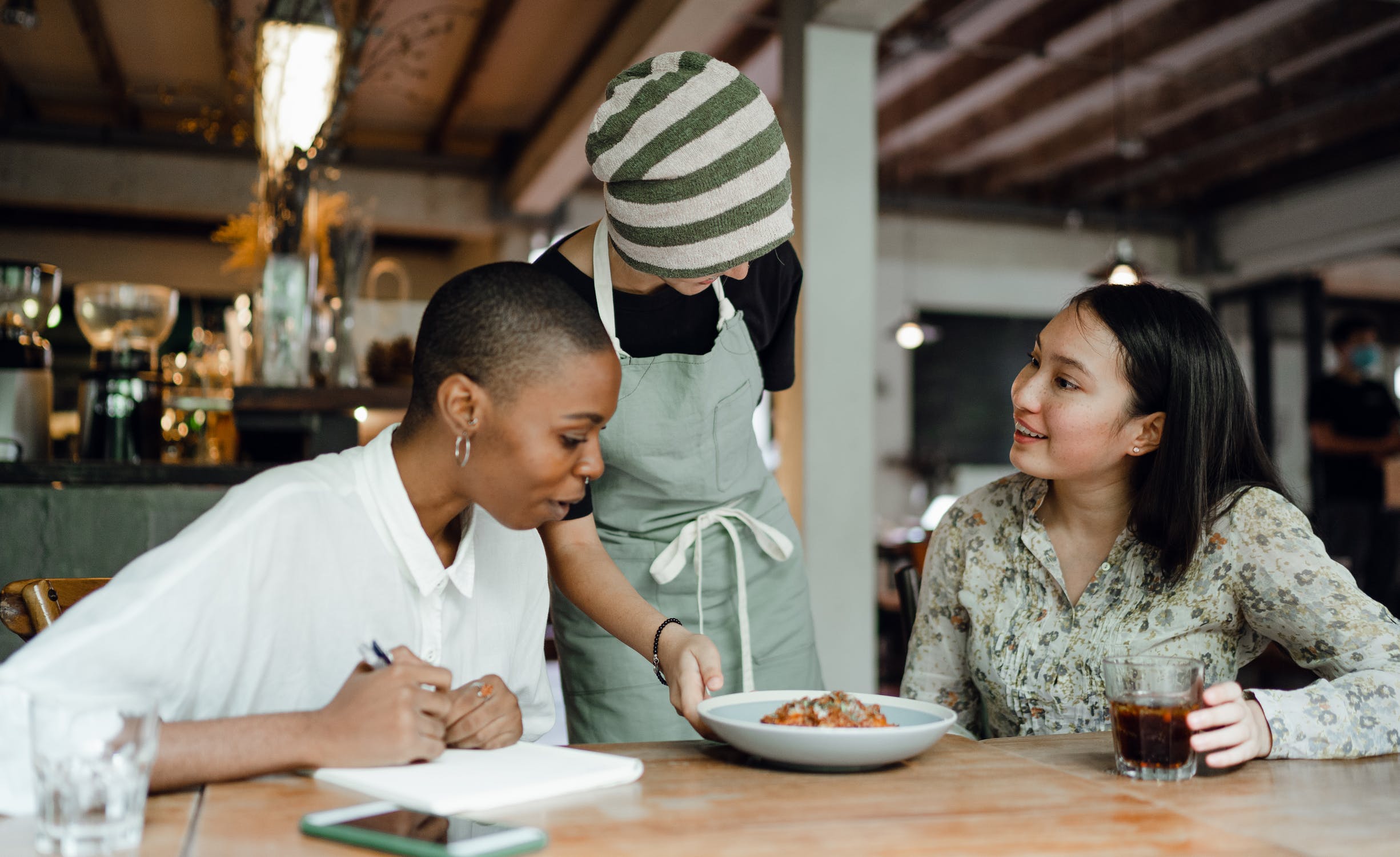 Two women sample a dish of food brought by a server. One is looking at the food, the other smiling at the server.