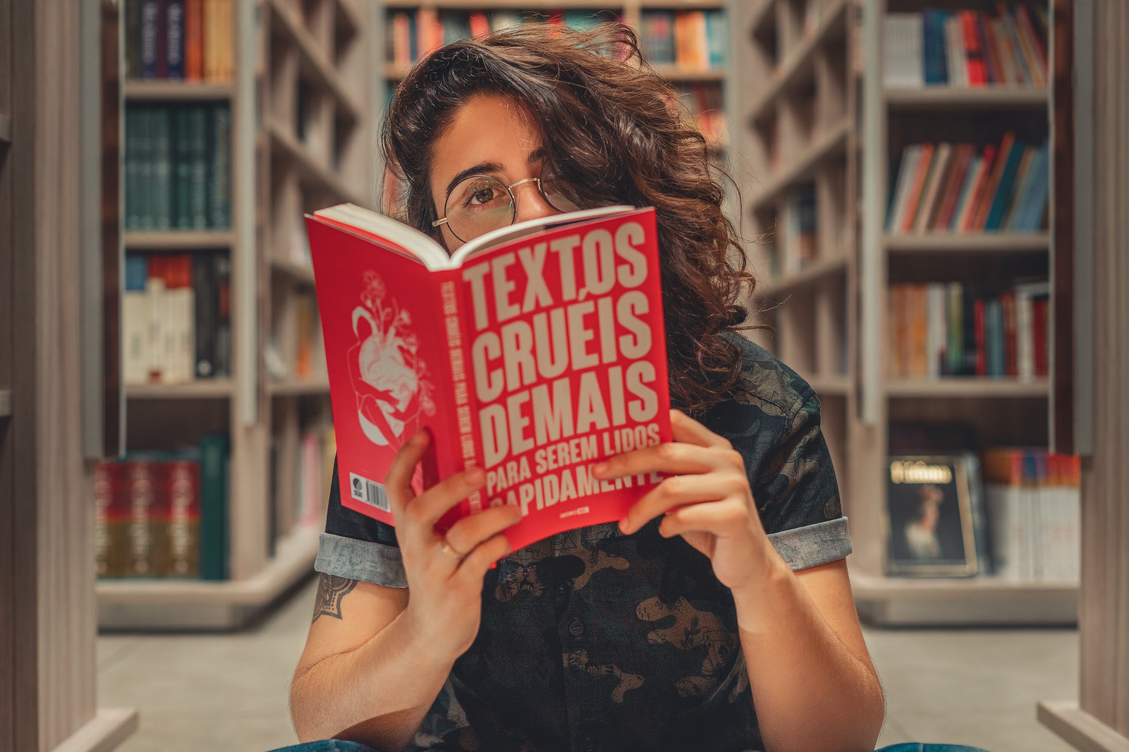 woman reading a book looking at the camera over the top of the book. the cover of the book reads, "text, os cures, demos para serum lidos."