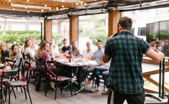 image of a man holding a microphone speaking to a group sitting a two long tables