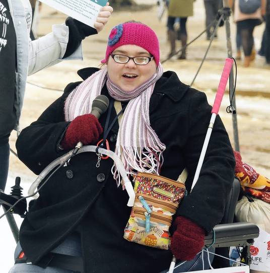 image of a woman wearing cold weather gear holding a microphone and a mobility walking stick, sitting in a wheel chair.
