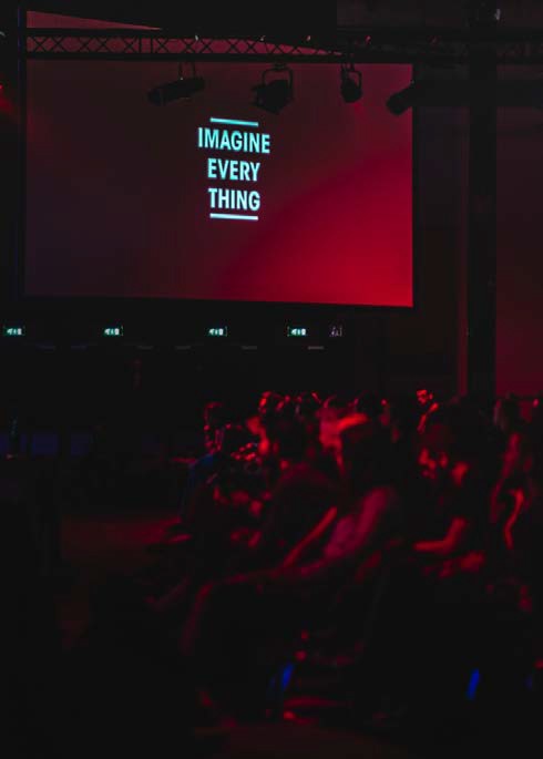 image of an audience in a red lit room looking at a project that reads "Imagine Every Thing"