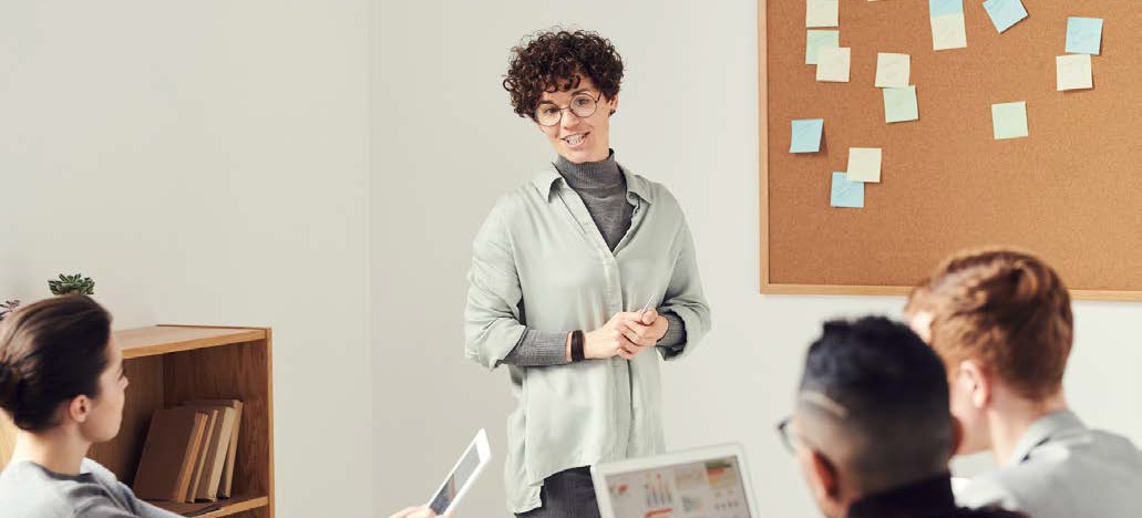 image of a woman standing in front of three people at a table. There is a corkboard with many sticky notes on it