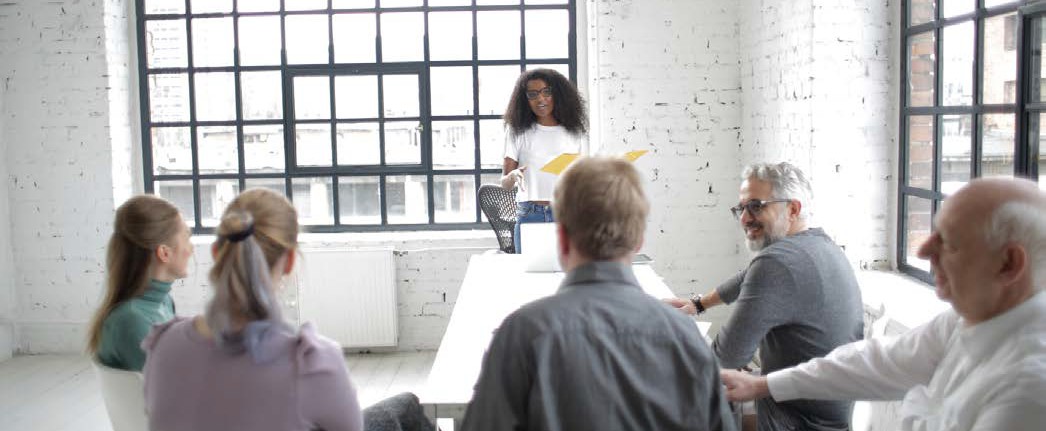 image of a woman standing in front of a group of five people sitting at a table in a bright white room with large windows