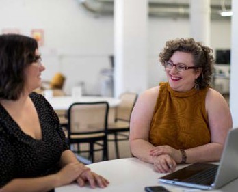 image of two women smiling at each other as they sit at a table in front of a laptop