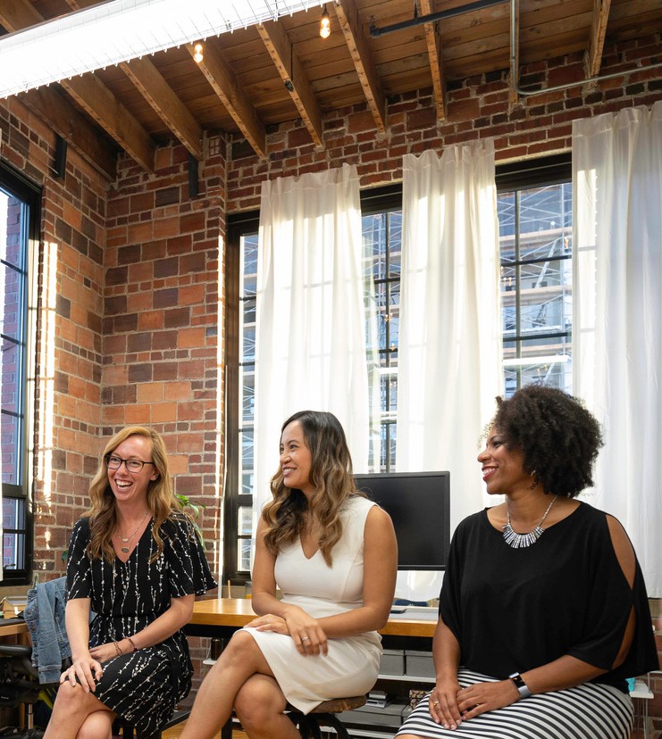 image of three women in business casual dress sitting in a row and smiling at something off screen. There is a bank of computer monitors behind them and floor to ceiling windows with white curtains