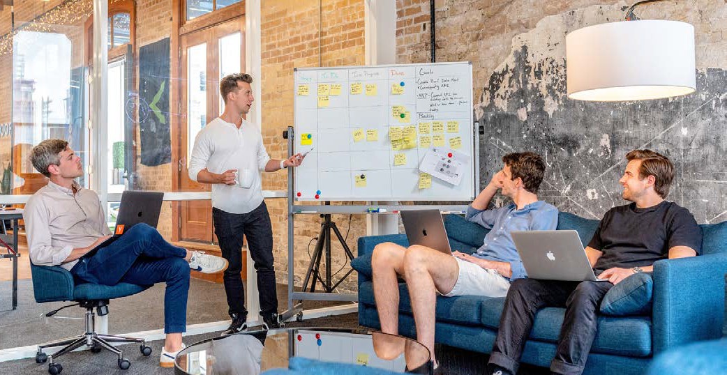 image of a man standing by a white board talking to a group of three other seated me holding laptops