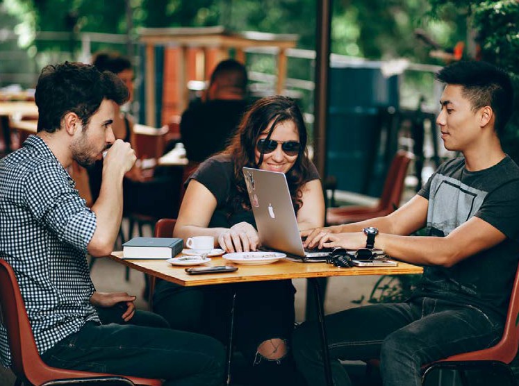 image of three people sitting at a table looking at a laptop together and drinking coffee