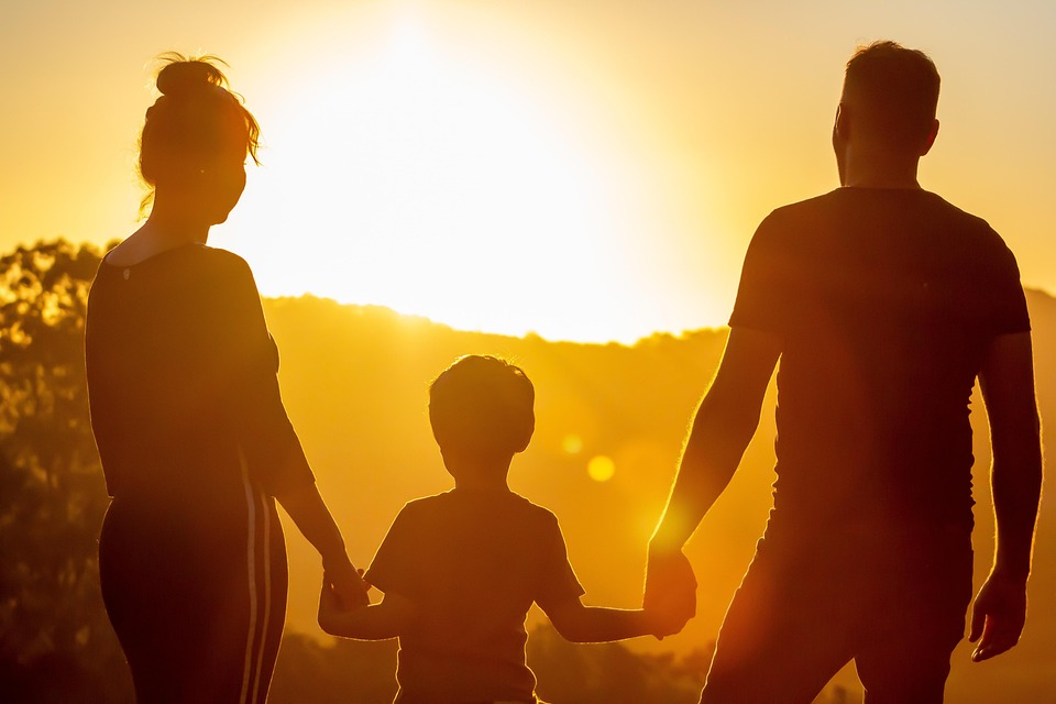 Image of a mother and father holding the hands of a child as the sun sets.