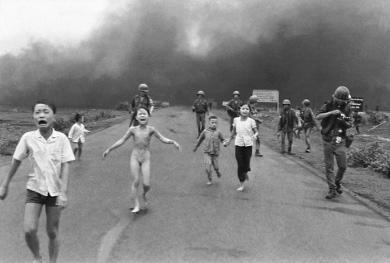 A photo of children running down a road with soldiers walking alongside in front of a dark cloud of smoke